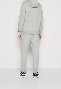 Persona che indossa un hoodie grigio chiaro e pantaloni della tuta abbinati, in piedi con le mani rilassate, scarpe Nike bianche e nere visibili.