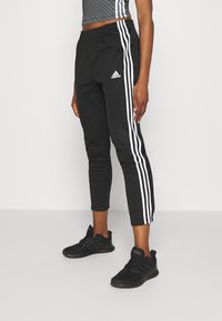 Svarta träningsbyxor med vita sidostråk, elastisk midja, cropped passform och tryckknappdetaljer längs sidan, tillsammans med svarta sneakers.