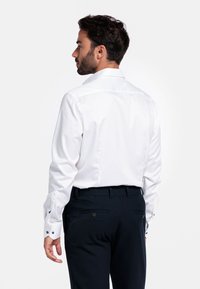 Witte overhemd met lange mouwen en een klassieke kraag, gecombineerd met donkerblauwe broek. Voorzien van knopen bij de manchetten en aan de achterkant.