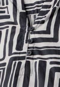 Camisa de patrones en blanco y negro con líneas geométricas, diseño de escote en forma de V y tres botones azules en la parte delantera. Textura de tela suave.