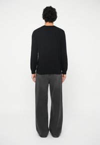 Άτομο με σγουρά μαύρα μαλλιά, ντυμένο με μαύρο μακρυμάνικο πουκάμισο και χαλαρούς σκοτεινούς γκρι τζιν, στέκεται στραμμένο πλάτη με φόντο λευκό.