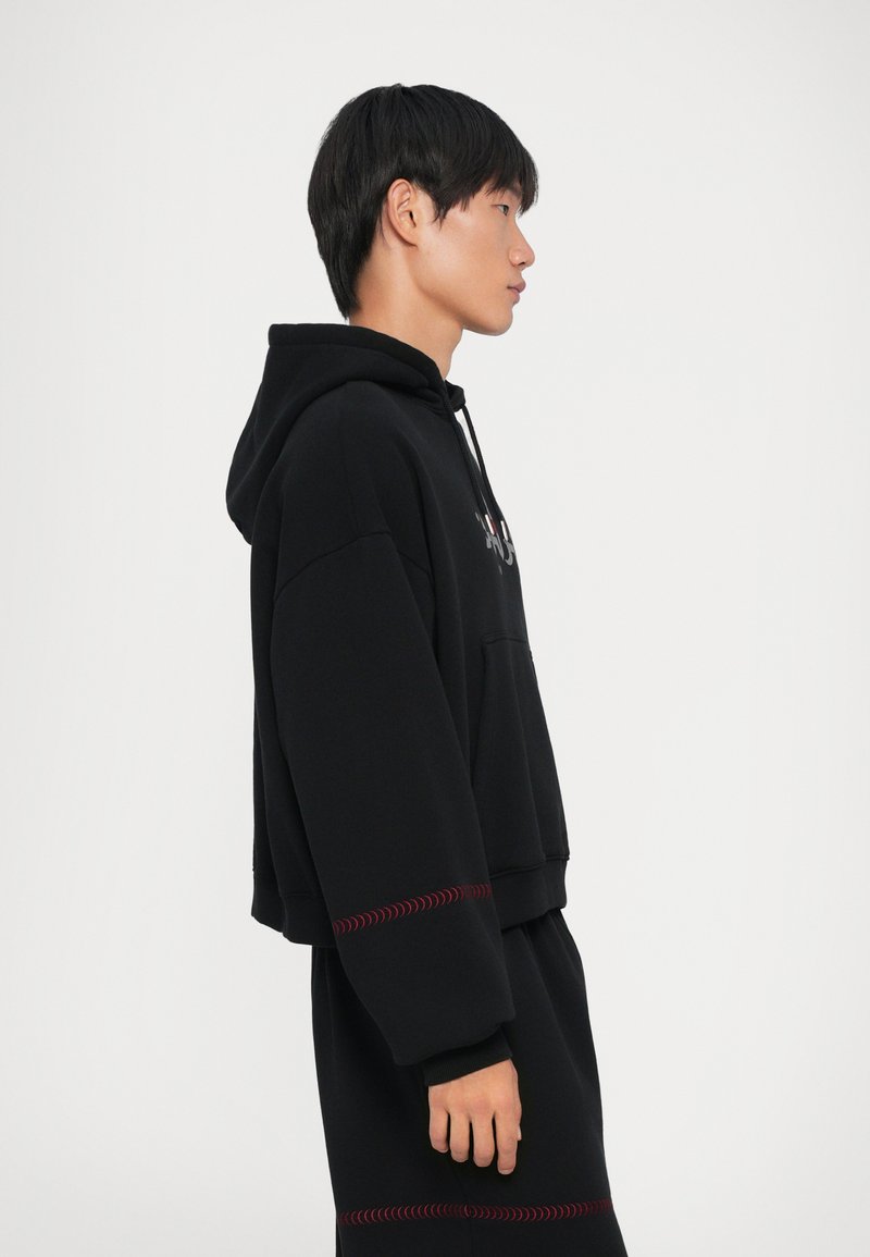 Sort cropped hoodie med en frontlomme, prydet med røde syninger ved ærmerne og kanten. Fremstillet af et blødt, struktureret stof.