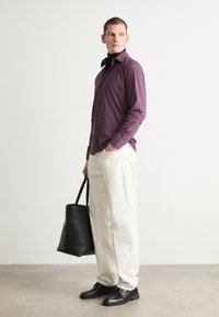 Paarse button-up shirt, witte broek, zwarte schoenen en een zwarte tote bag. Model staat met één hand in de zak en laat een strakke ontwerp zien.