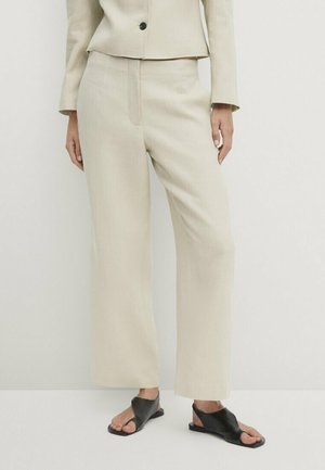 Kvinde iført beige bukser med vide ben og matchende blazer med sorte åbne flade sandaler, stående mod en ensfarvet baggrund.