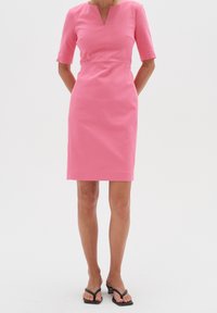 Rosa åtsittande klänning med korta ärmar, V-halsringning och sömdetaljer, som bärs med svarta flip-flops. Slät textur, knälång design.