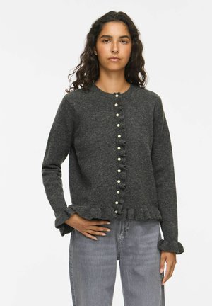 Vrouw met lang donker krullend haar draagt een donkergrijs vest met knopen en ruches aan de manchetten en zoom, gecombineerd met lichtgrijze spijkerbroek.