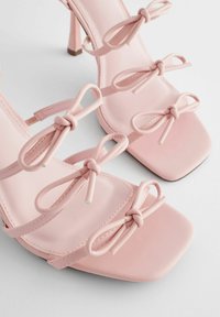 Hellrosa Absatzsandalen mit quadratischen Zehen und drei dünnen Riemen, die in kleinen Schleifen über den Fuß gebunden sind.