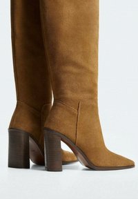 Bottes en daim marron, arrivant au genou, avec un bout pointu et un talon épais, présentant une texture lisse et un design minimaliste.