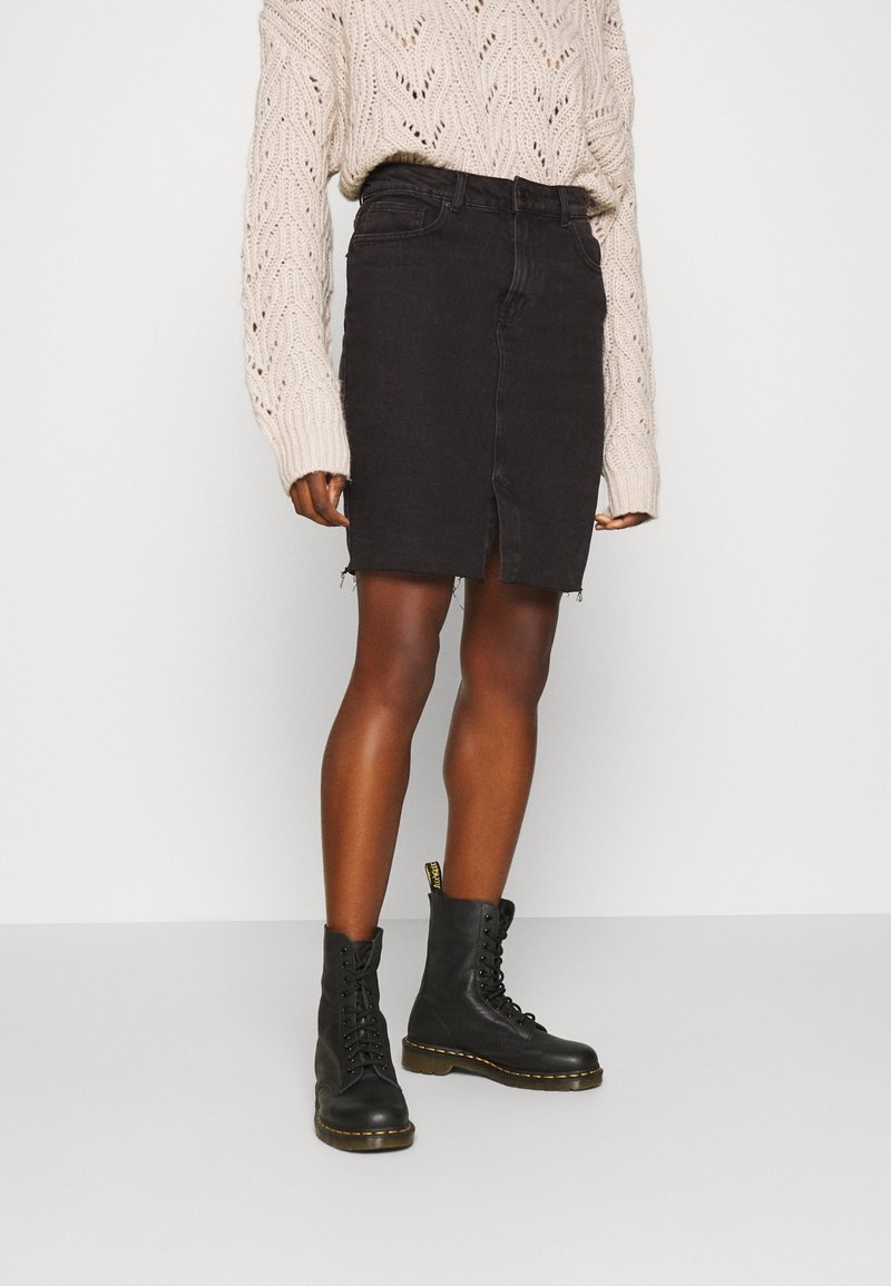 Falda de mezclilla negra con una abertura frontal, combinada con un suéter de punto grueso en color crema y botas negras con cordones. El conjunto destaca por el contraste de texturas.