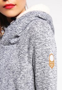 Harmaa, neulottu huppari, jossa on pehmeä, valkoinen fleecevuori. Sisältää sivutunnisteen, jossa on logo. Kankaassa on rakenteellinen kuvio.