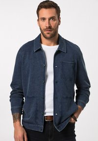 Tmavě modrá bunda vyrobená z džínoviny s límcem, zapínáním na knoflíky a náprsní kapsou. Obsahuje volný střih a dlouhé rukávy.