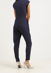 Marinblå jumpsuit med korta ärmar, figursydd midja och avsmalnande ben. Silverfärgade högklackade skor med ankelremmar kompletterar outfiten.