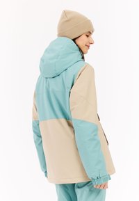 Leichte, zwei-farbige Jacke in Beige und Blaugrün. Mit Kapuze, elastischen Bündchen und glatter Textur. Getragen mit einer beigen Strickmütze.