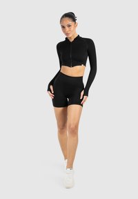 Sort cropped zip-up jakke med lange ærmer, parret med højhvide sorte shorts. Glat tekstur, tætsiddende design og minimalistisk stil.