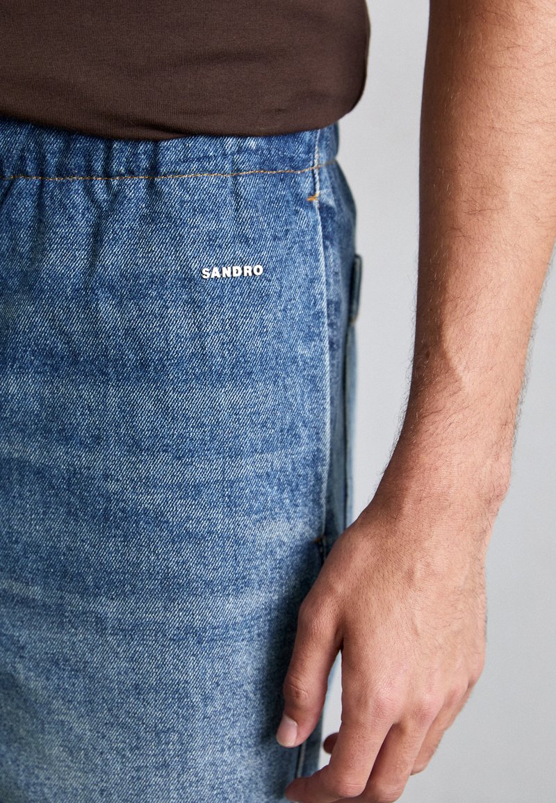 Pantaloni in denim blu chiaro con cuciture visibili e una cintura elastica. Logo "SANDRO" in bianco sulla parte anteriore. Mano appoggiata di lato.