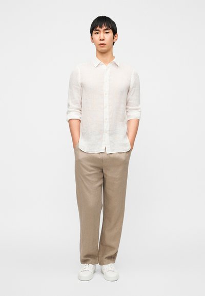 Λευκό λινό πουκάμισο με κουμπιά, μανίκια γυρισμένα, ελαφρύ υφή. Μπεζ λινό παντελόνι, άνετη εφαρμογή. Λευκά αθλητικά παπούτσια, ουδέτερο σχέδιο.