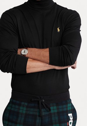 Uomo che indossa una maglia nera a maniche lunghe con logo ricamato giallo, pantaloni a quadri verdi e blu con coulisse e un orologio da polso, con le braccia incrociate.