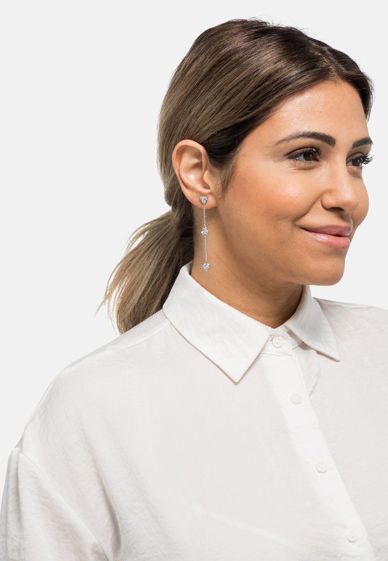 Lunghi orecchini d'argento con tre accenti di gemme trasparenti, caratterizzati da un design di catena delicato. Il modello indossa una camicia bianca con colletto.