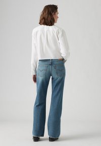 Jeans en denim bleu clair évasés avec une taille mi-haute, des coutures visibles et deux poches arrière. Associés à une chemise à manches longues blanche.