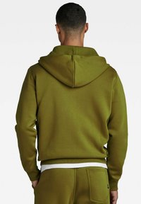 Felpa con cappuccio verde oliva dal tessuto liscio, con polsini a coste e tasca a marsupio. Presenta un bordo bianco all'orlo e un design semplice.
