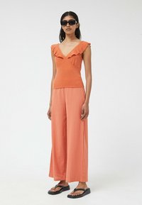 Top naranja con volantes y escote en V, combinado con pantalones anchos texturizados en un tono a juego. La modelo lleva gafas de sol negras y chancletas.
