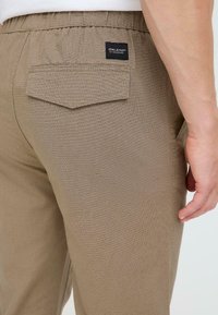 Beige textured pants with elastic waistband, back pocket, and branding label. Smooth fabric with subtle pattern and straight cut.