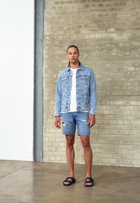 Helehall denimíjak, valge T-särgi peal, koos kulunud denimilühikeste pükste ja mustade sandaalidega. Riietust täiendab sile betoontaust.