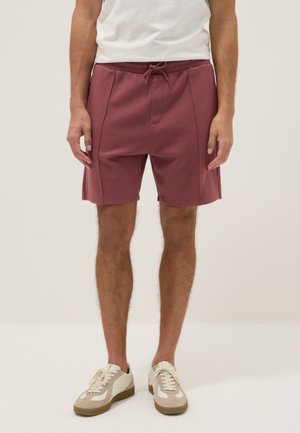 Uomo che indossa pantaloncini bordeaux con coulisse, t-shirt bianca e sneakers bianche beige, in piedi su uno sfondo chiaro uniforme.