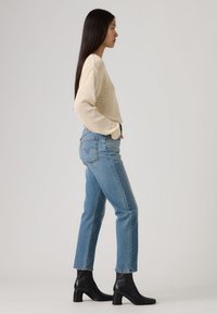 Μπεζ πουλόβερ με υφή, ανοιχτά μπλε τζιν με πίσω τσέπες και μαύρα δερμάτινα μποτάκια αστραγάλου με φαρδιές τακούνια, εμφανίζονται από το πλάι.