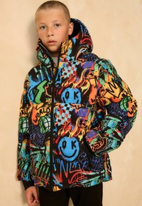 Un garçon portant une veste matelassée à capuche avec un motif de graffiti coloré, une fermeture éclair et un design de smiley, se tenant devant un fond beige.