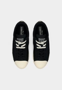 Ζευγάρι μαύρα αθλητικά παπούτσια Converse All Star με λευκές λαστιχένιες μύτες και ασύμμετρα κορδόνια λευκού και μαύρου χρώματος σε λευκό φόντο.