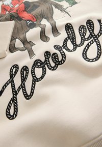Krämfärgad tyg med en tryckt bild av en man i röd färg på en häst, med svart rep-liknande broderad text som säger "Howdy."