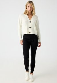 Cárdigan blanco de punto con cuello en V, tres botones grandes y bolsillos delanteros, combinado con leggings negros y zapatillas blancas.