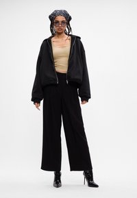 Crna oversized zip-jakna, bež skraćeni top, crne široke hlače i crne cipele s ravnim potpeticama; nosi uzorčanu maramu i sunčane naočale.