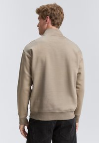 Beige pulóver magas gallérral, bordás mandzsettákkal és aljjal, hátulról nézve. Simára szőtt anyag; minimalista dizájn. Fekete farmer látható.
