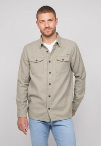 Chemise à manches longues gris clair avec fermeture à boutons, deux poches poitrine et un col. Portée par-dessus un t-shirt blanc, associée à un jean bleu.