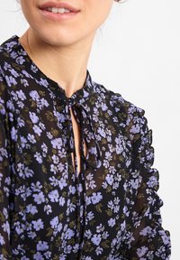 Schwarze Bluse mit floralen Mustern in Lila und Grün, mit Rüschensäumen, Bindeband am Hals, leichtem Stoff und einer leicht durchsichtigen Textur.