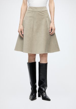 Femme portant une jupe beige trapèze avec une texture diagonale et des bottes noires hautes à motif crocodile, debout devant un fond uni.