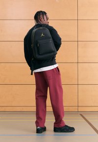 Svart ryggsekk med gull aksenter og frontlomme, båret over en svart genser og burgunder joggebukser, kombinert med svarte joggesko.