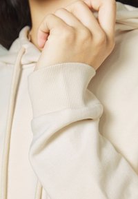 Sweat-shirt beige avec un poignet côtelé. Le tissu semble doux avec une texture lisse, présentant un design simple et décontracté.