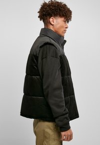 Vesta puffer neagră cu textură de catifea pe jumătatea inferioară. Guler înalt și închidere cu fermoar. Poartă peste un tricou negru cu mânecă lungă, stilizată cu pantaloni kaki.