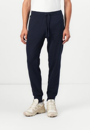 Navy blue jogger pants with elastic waistband and drawstring. Features side zippers and tapered legs. Paird with light beige athletic shoes.