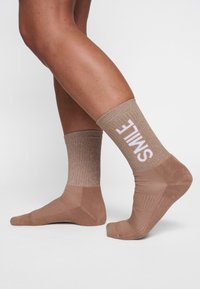 Chaussettes marron avec une partie supérieure texturée et scintillante et une partie inférieure unie. Texte blanc "SMILE" sur le bracelet, mettant en valeur le design.
