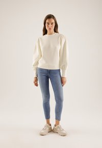 Vrouw met een crèmekleurige top met lange mouwen en ballonvormige schouders, blauwe strakke jeans en beige sneakers, staand tegen een effen achtergrond.