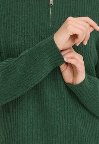 Zöld csíkos pulóver negyed cipzárral. Az anyag texturált; az ujjak szűkített mandzsettával rendelkeznek. A kezek az ujját igazítják.