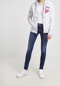 Sudadera gris con cremallera y detalles gráficos rojos, llevada sobre una camiseta blanca, combinada con pantalones vaqueros azul oscuro y zapatillas blancas.