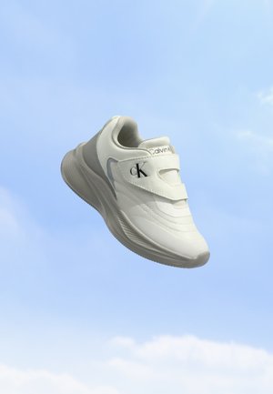 Hvid Calvin Klein sneaker med velcroremme, der svæver mod en blå himmelbaggrund.