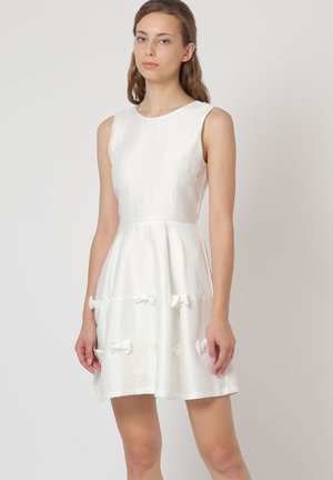 Vestido blanco sin mangas con un cuerpo ajustado y falda acampanada, que presenta pequeños lazos de tela a lo largo del dobladillo. Hecho de un material texturizado.