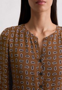 Braune Bluse mit dezentem Muster aus weißen und roten Tropfenformen, Rundhalsausschnitt und Knopfdetails vorne. Glatte Stoffoberfläche.