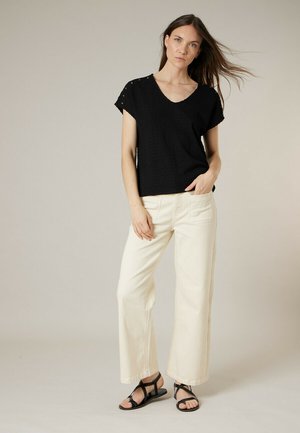 Vrouw met lang bruin haar, gekleed in een zwart kortärmelig topje, crèmekleurige broek met wijde pijpen en zwarte sandalen met bandjes, staand tegen een neutrale achtergrond.